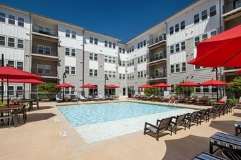 A swimming pool with chaise lounge chairs and umbrellas in front of an apartment building